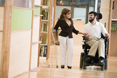 A woman walking down a hall with a man in a powered chair